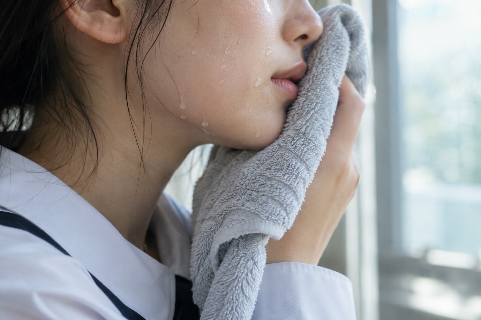 창가에서 흰 교복 셔츠 차림 학생이 물방울 맺힌 볼과 입가에 회색 수건을 대고 있는 옆얼굴