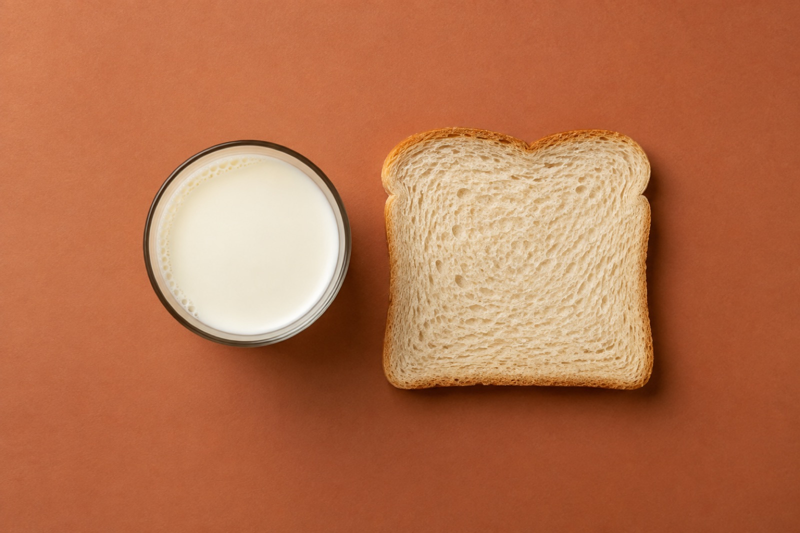 A glass of milk and a slice of white bread side by side, shot from above on a terracotta background