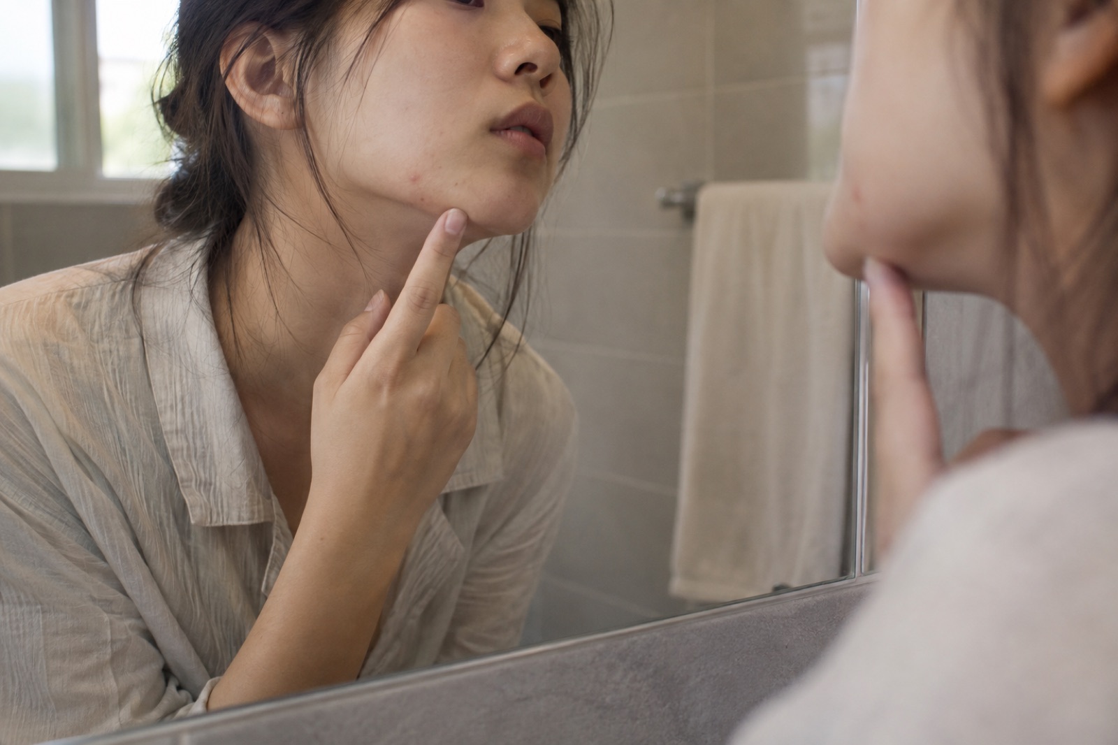 浴室の鏡に映る女性が、人さし指をあごの先に当てて、ほおの小さな赤い点をひとつのぞき込んでいる様子