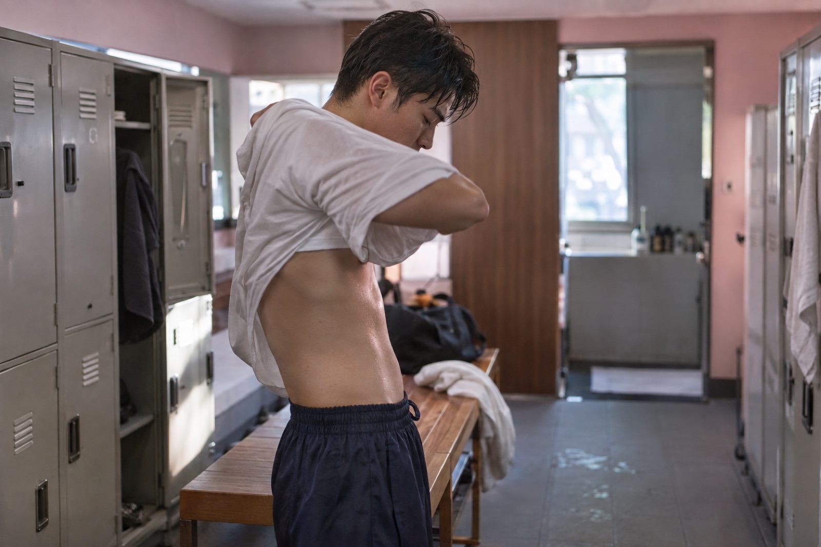 A man pulling a sweat-soaked white T-shirt over his head in front of gray lockers in a locker room with pink walls