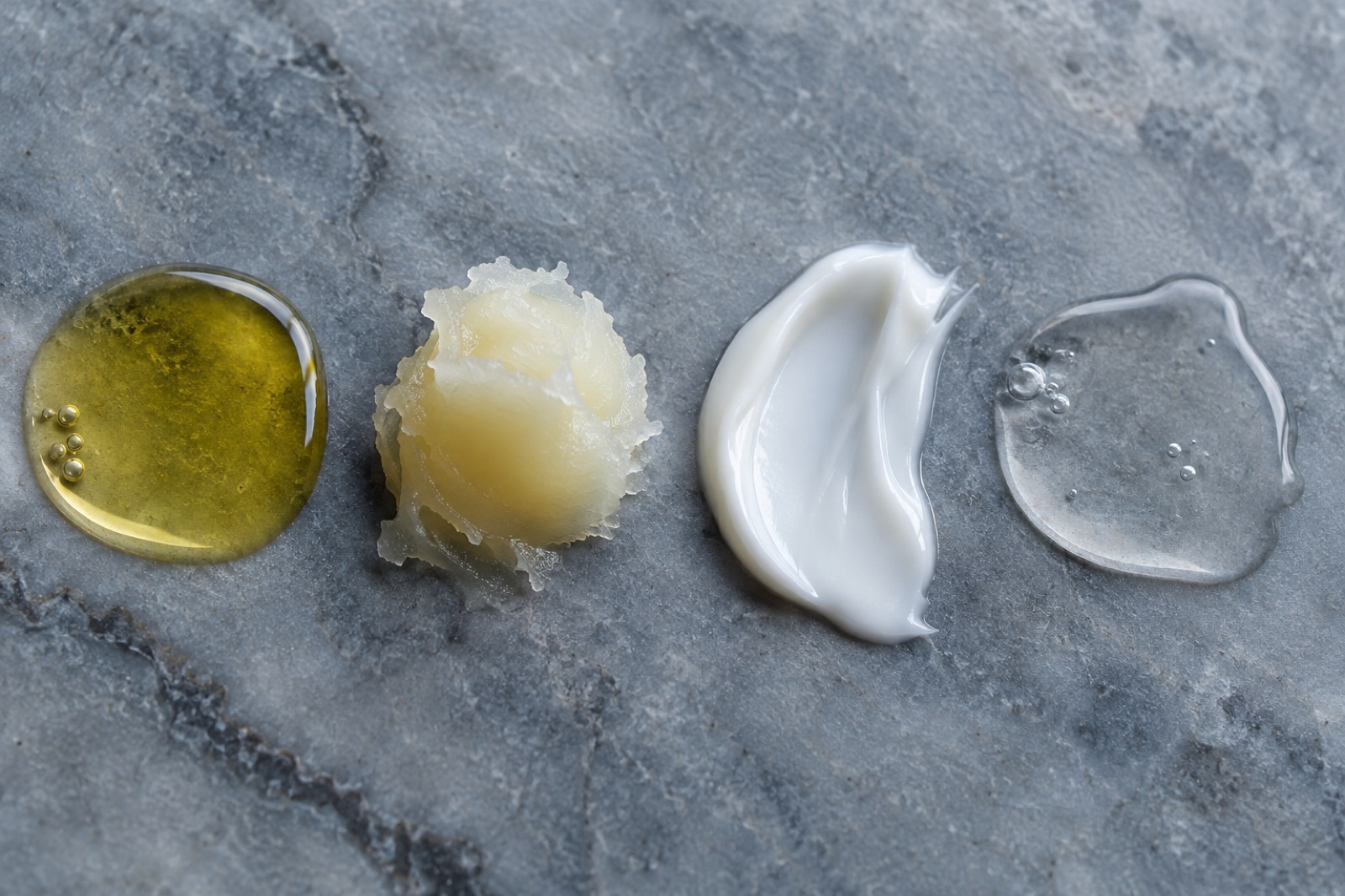 ออยล์สีเหลือง บาล์มสีเหลืองอ่อนเป็นก้อน ครีมสีขาว และของเหลวใสมีฟองอากาศ วางเรียงจากซ้ายไปขวาบนหินอ่อนสีเทา