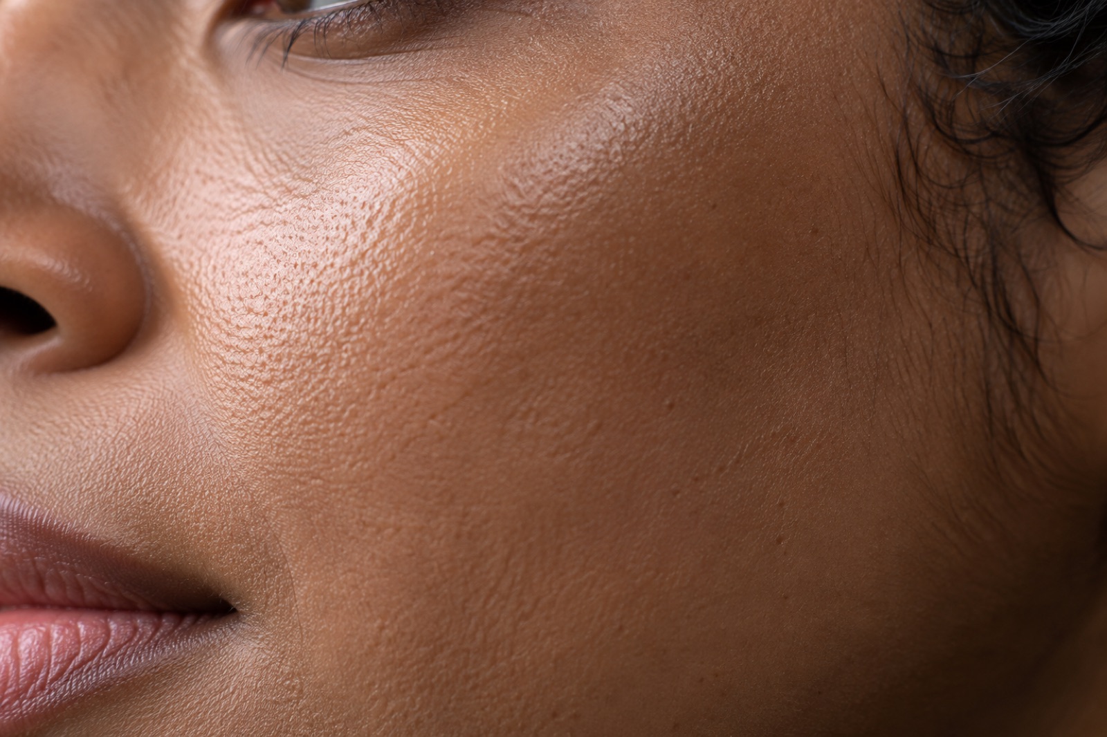 Close-up of fine texture and pores on brown-toned cheek skin running toward the side of the nose and the corner of the lips