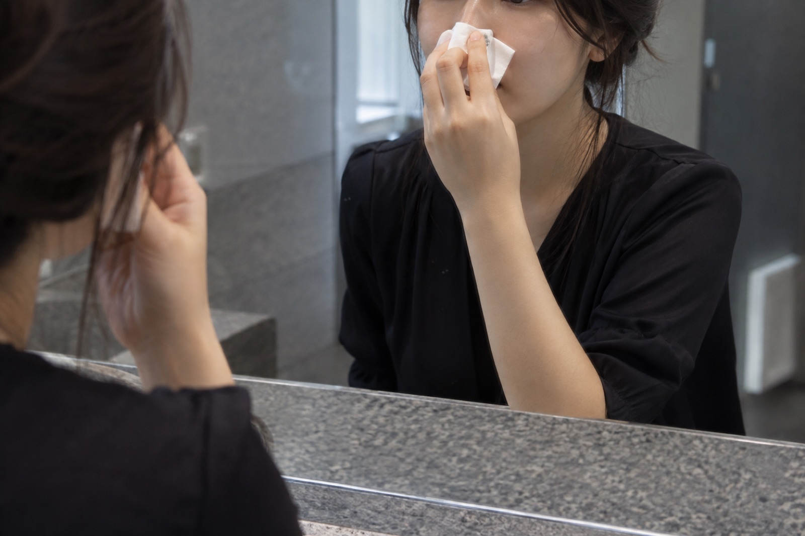A woman in a black blouse, reflected in a mirror above a gray stone sink, pressing a white tissue against the bridge of her nose