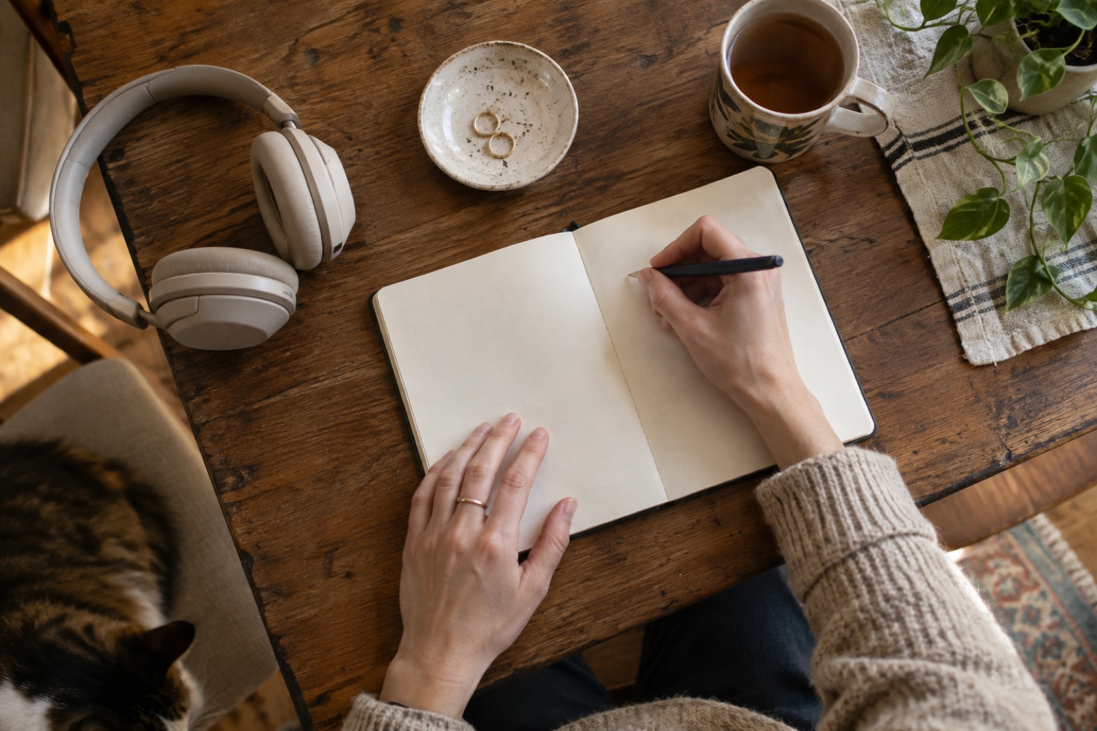 Looking down at a wooden table with beige headphones, a small dish holding two gold hoops, and a teacup, while a hand holds a pen to a blank notebook