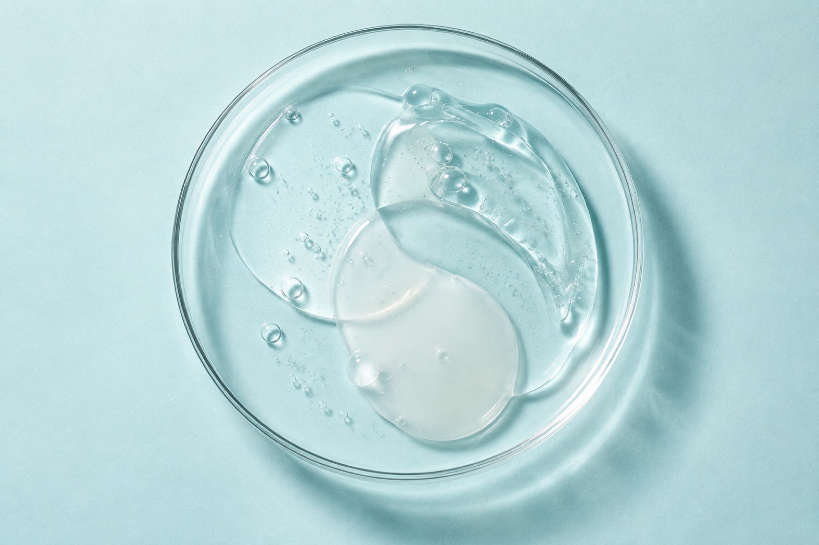Gel trong suốt và dung dịch trong mờ loang ra chồng lên nhau trong chiếc đĩa petri thủy tinh tròn trên nền xanh da trời