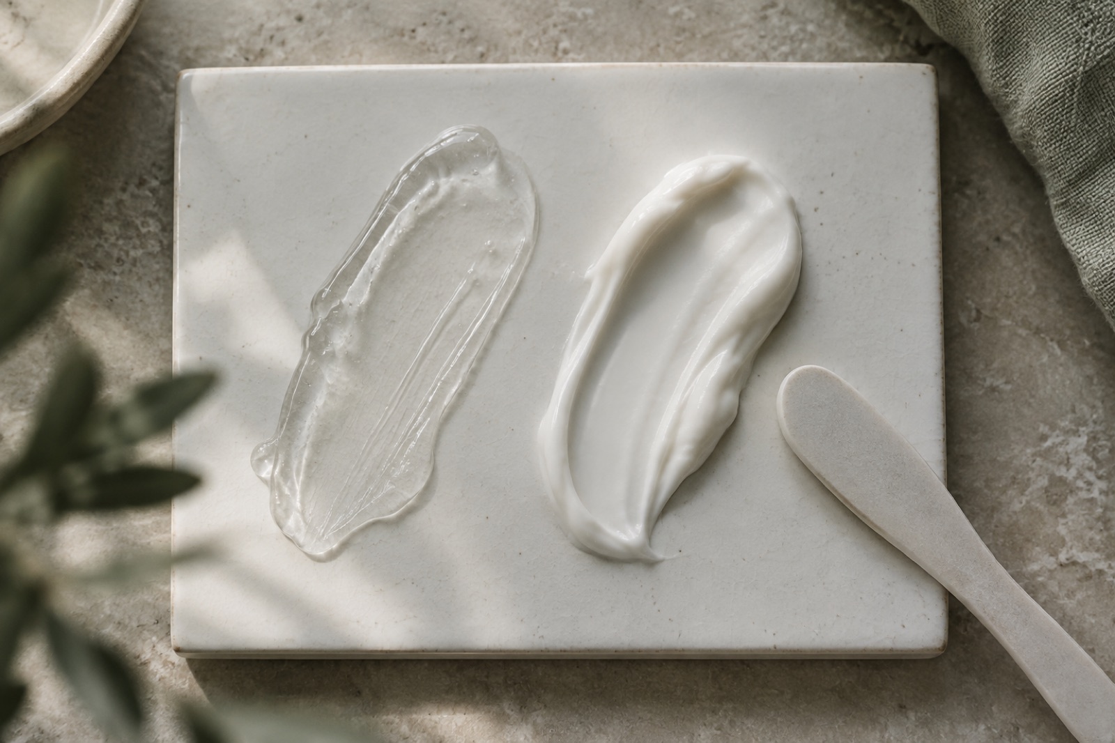 Top view of clear gel on the left and white cream on the right spread in long strokes on a white tile over beige stone, with a spatula