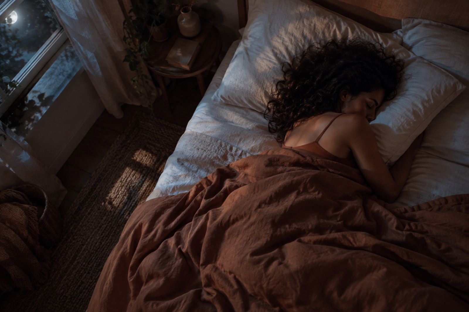 A curly-haired woman asleep almost face-down with one cheek buried in a white pillow, the moon visible through the bedroom window