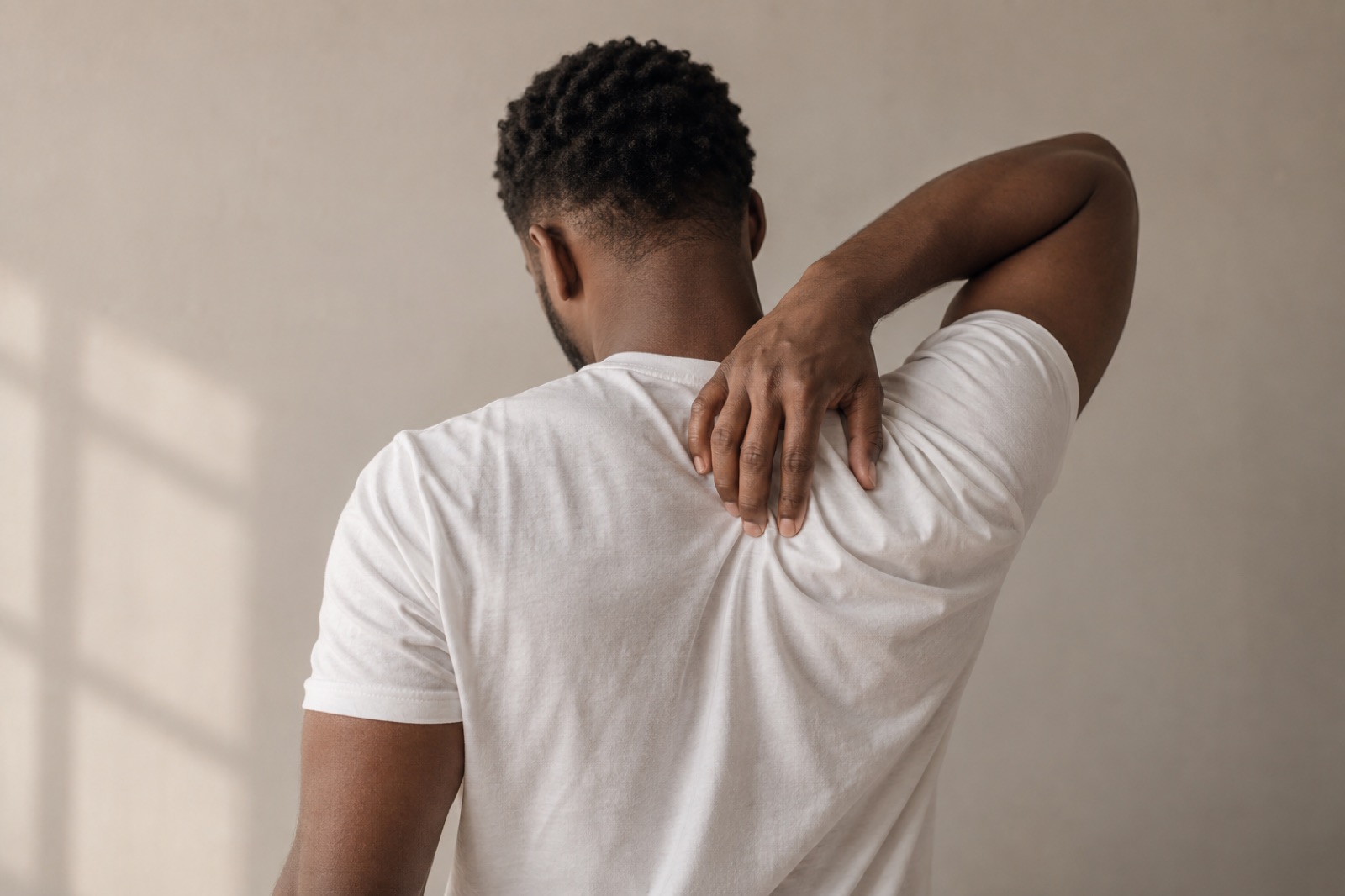 Back view of a man in a white short-sleeve T-shirt against a beige wall, reaching one hand over his shoulder to his upper back