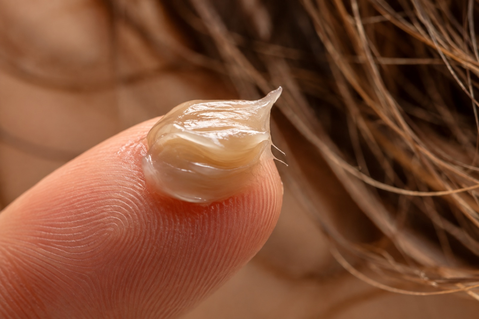 Close-up of a translucent cream-style hair product on the tip of an index finger in front of tousled hair