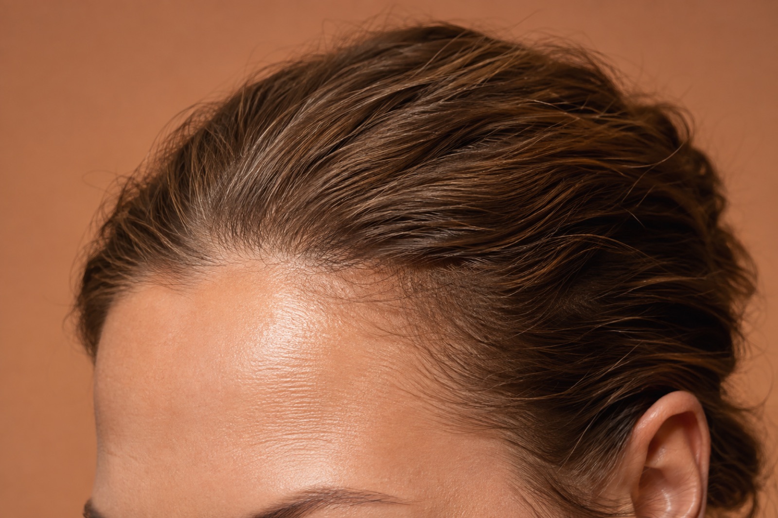 Close-up against an orange background of brown hair combed back, showing the front hairline and the top of the ear