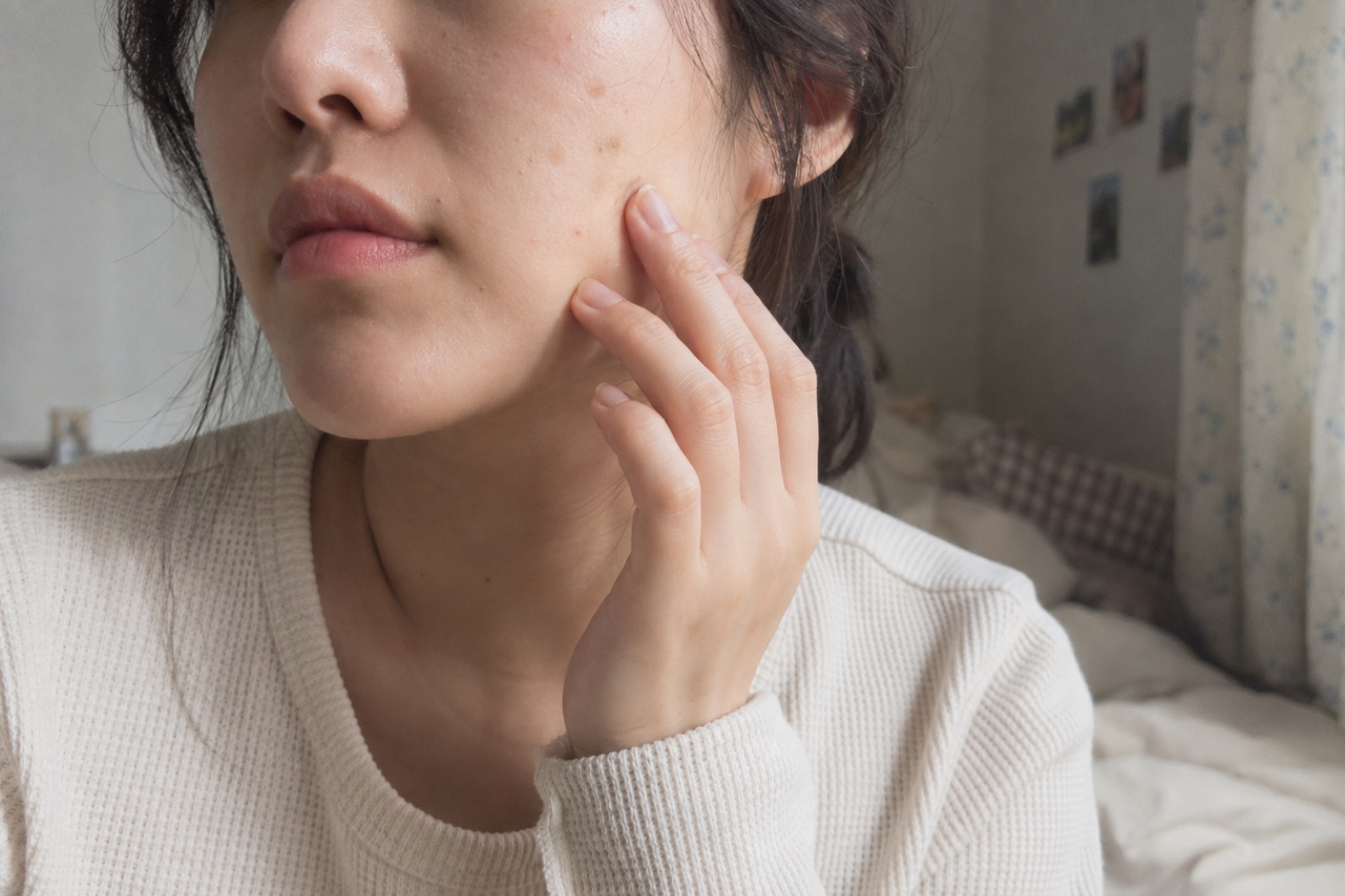 A woman in a white waffle-knit top rests her fingertips on her cheek above the jawline, with a few faint brown spots on the cheek beside her nostril