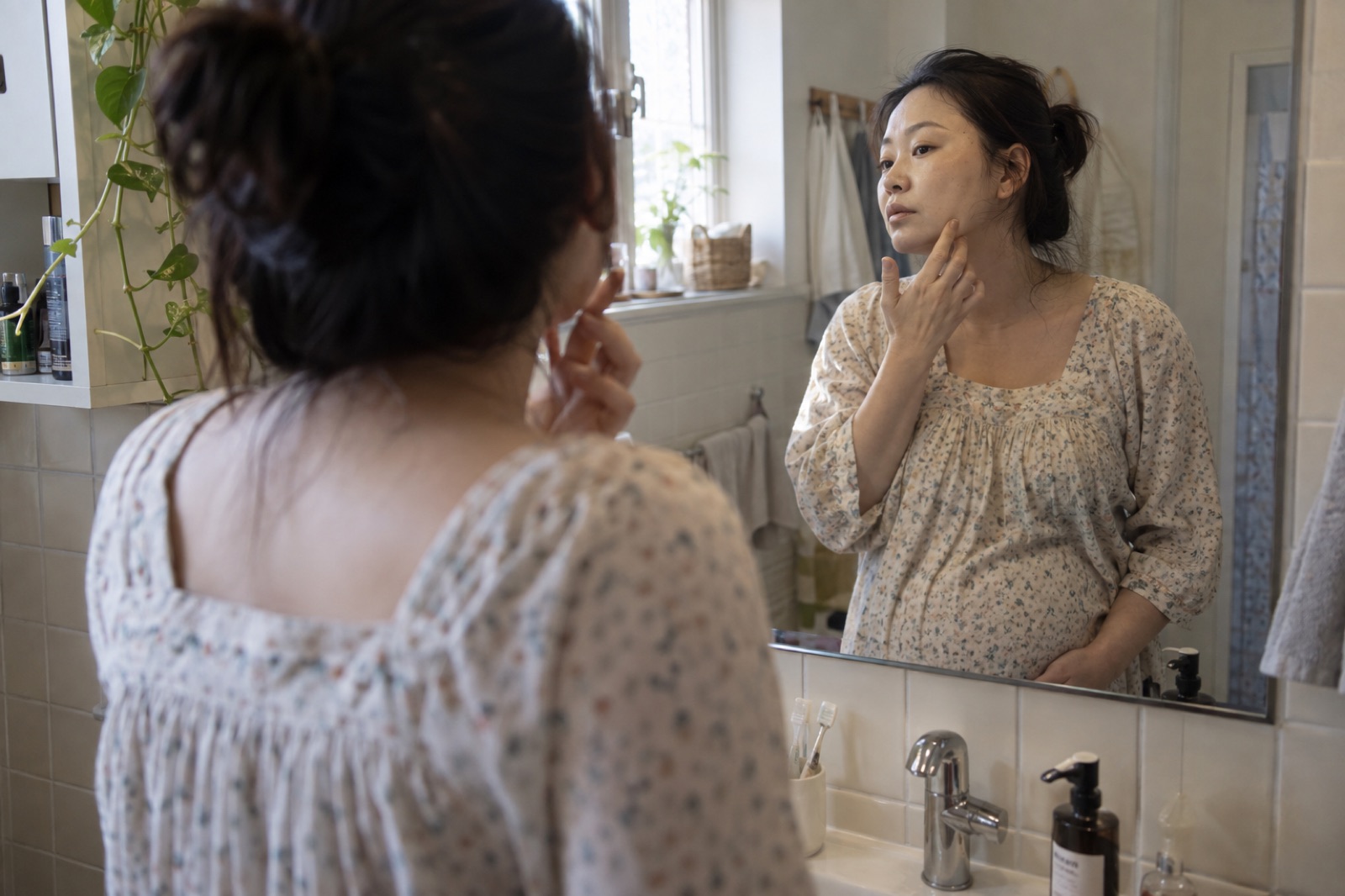 小花柄のワンピースを着た妊婦が洗面台の鏡に顔を映し、2本の指で口元の横の頬にふれている後ろ姿