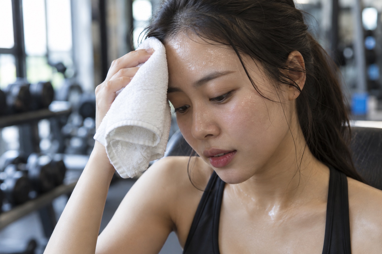 Người phụ nữ lấm tấm mồ hôi cúi đầu, áp chiếc khăn trắng lên trán trong phòng tập có dụng cụ mờ phía sau