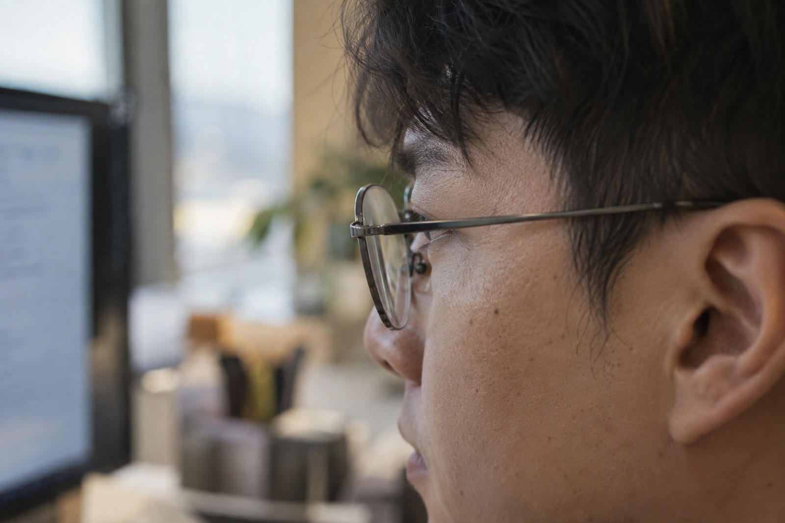 Side view of a man in thin metal-framed glasses at a monitor, the glasses arm running from the corner of his eye to above his ear